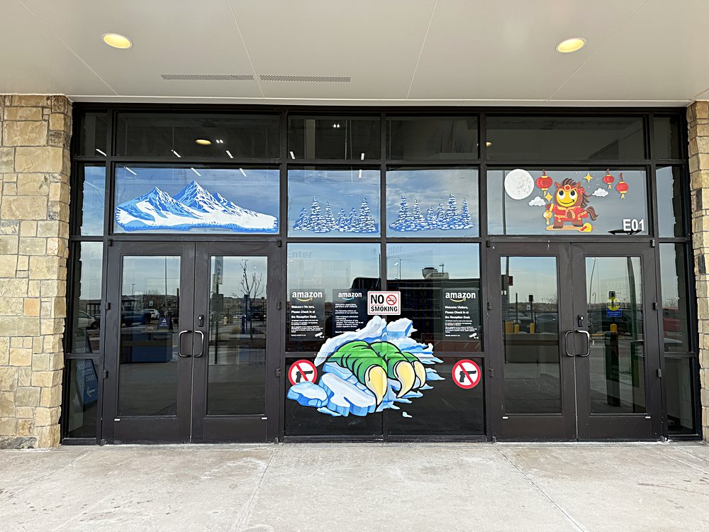 Hand-painted seasonal window display on the entrance glazing of an Amazon fulfillment center in Colorado, february 2026. snow peaks, and the site mascot mid-slide