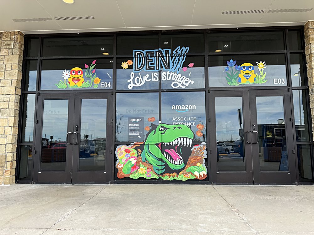 Hand-painted seasonal window display on the entrance glazing of an Amazon fulfillment center in Colorado, march 2026. mascot and blossom across the top band