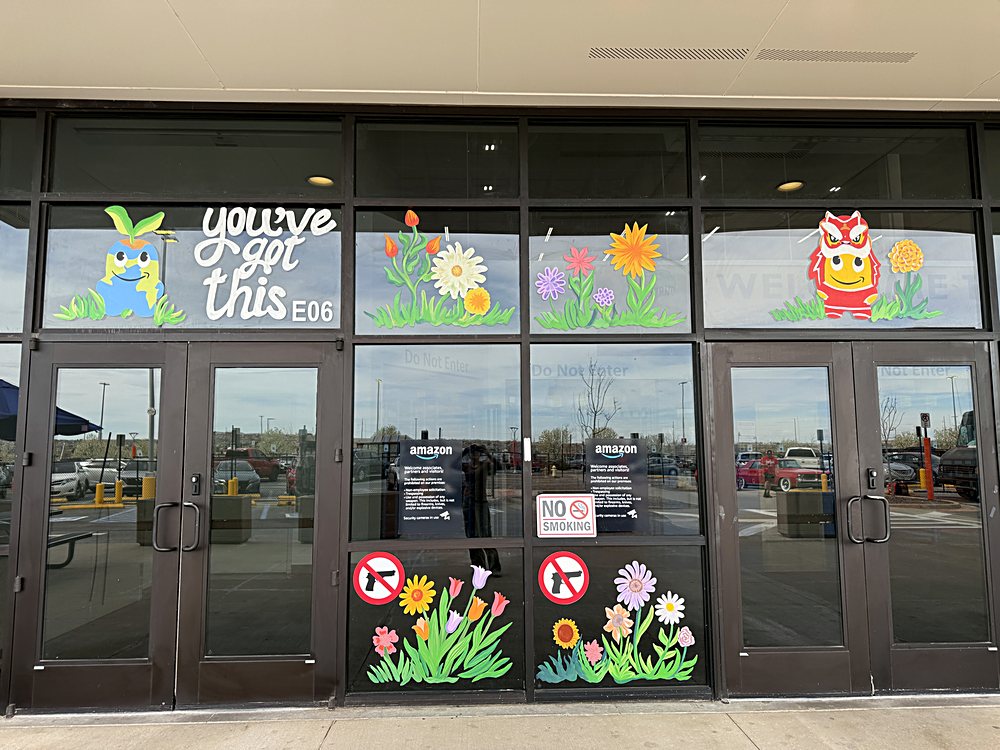 Hand-painted seasonal window display on the entrance glazing of an Amazon fulfillment center in Colorado, march 2026. lettering and spring characters at door height