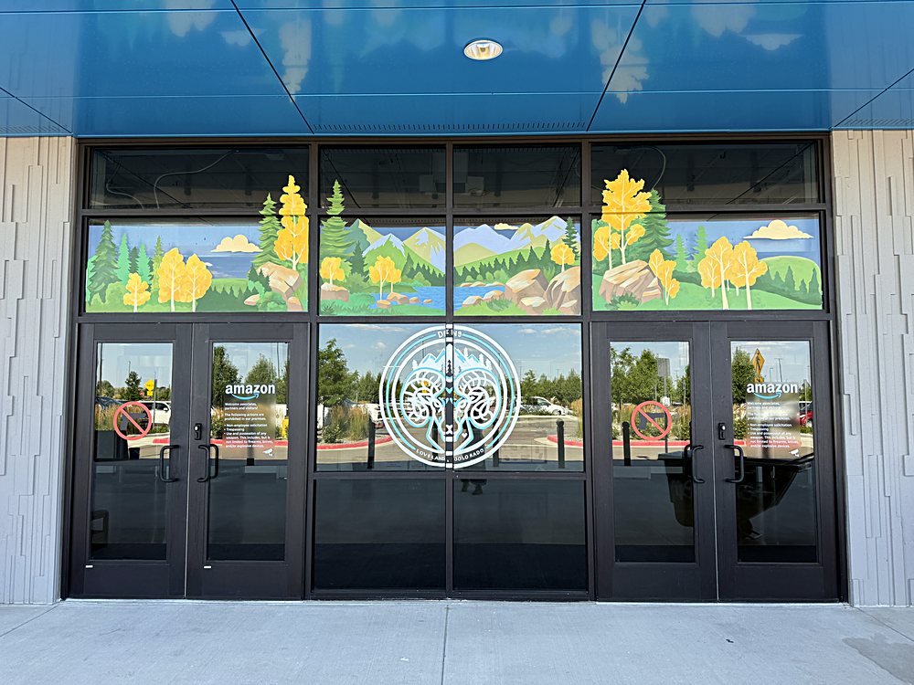 Hand-painted seasonal window display on the entrance glazing of an Amazon fulfillment center in Colorado, july 2026. aspen turning, across four panels