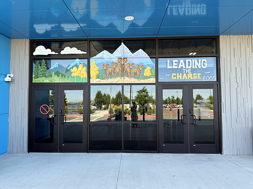 Hand-painted seasonal window display on the entrance glazing of an Amazon fulfillment center in Colorado, july 2026. peaks and pines above the doors